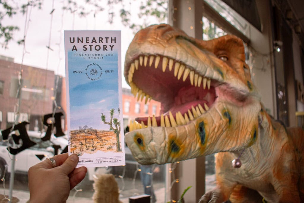 hand holding up a summer adventure bookmark in front of a dinosaur